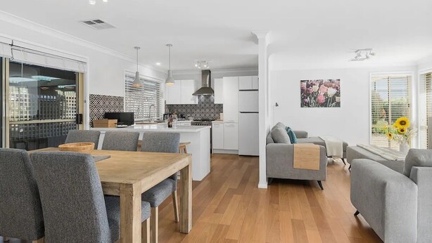 Una cocina moderna con armarios blancos, una isla central e iluminación colgante. Hay un comedor con sillas tapizadas de color gris y una mesa de madera. Un sofá gris y sillones están ubicados cerca de un ventanal con vistas al exterior.
