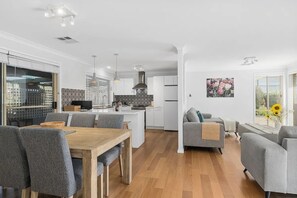 A modern kitchen with white cabinets, a central island, and pendant lighting. There is a dining area with grey upholstered chairs and a wooden table. A grey sofa and armchairs are positioned near a large window with a view of the outdoors. - The Scotty House - EV and pet friendly (Moss Vale)