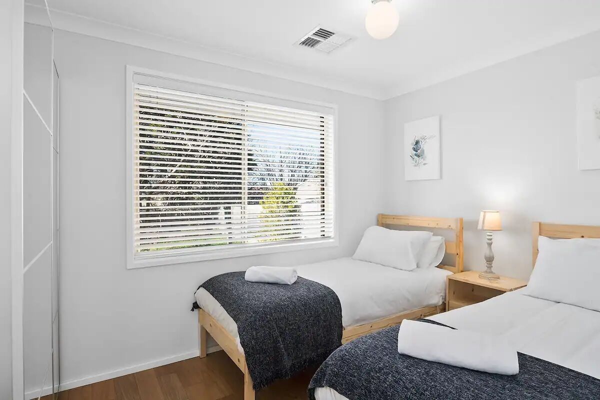 A bedroom with two single beds, a large window with blinds, a bedside table with a lamp, and a framed picture on the wall.