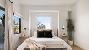Une chambre à coucher moderne avec un grand lit, des tables de chevet, un banc et une vue sur l’extérieur.