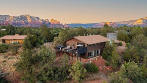Une maison moderne à deux étages, dotée d’une terrasse et d’un espace salon extérieur, entourée d’une forêt dense et avec des montagnes en toile de fond.