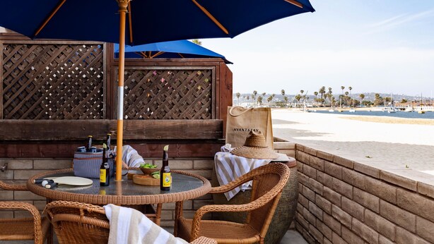 An outdoor patio with wicker furniture, a table with a bottle of beer, and a bag labeled "AVANTSTAY". - Patio + Sunset Views + Beach Days | Right on the Water | Bay View 2 (San Diego)