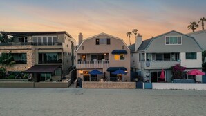 A beachfront property with a two-story house featuring a balcony, a covered patio with umbrellas, and a beach view. - Patio + Sunset Views + Beach Days | Right on the Water | Bay View 2 (San Diego)