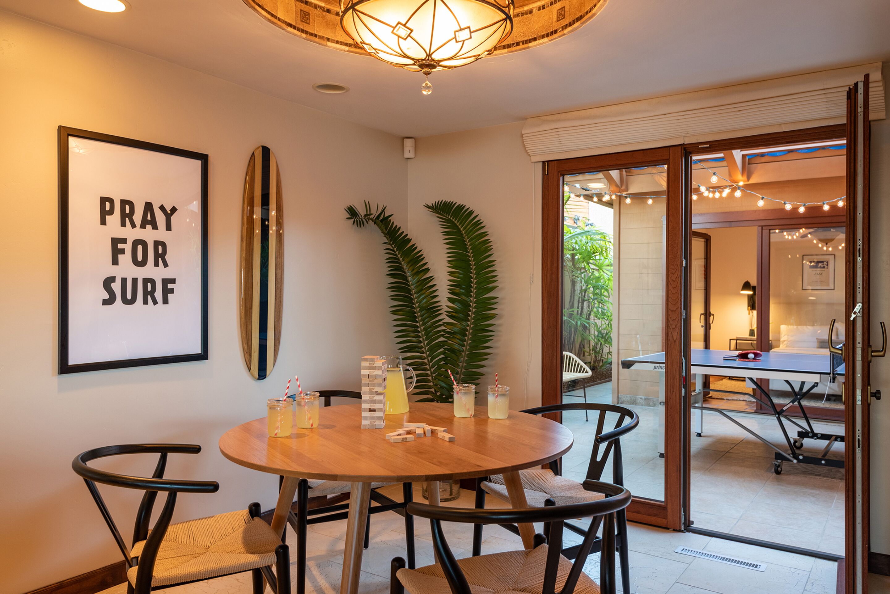A dining area with a round table, chairs, a surfboard, a framed sign, and a plant.
