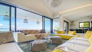 Un salon moderne avec un canapé, des poufs et un lit jaune, de grandes fenêtres donnant sur la mer et des lampes suspendues.