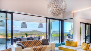 Un salon moderne avec un canapé, des fauteuils et un coin repas. De grandes fenêtres offrent une vue sur l’extérieur.
