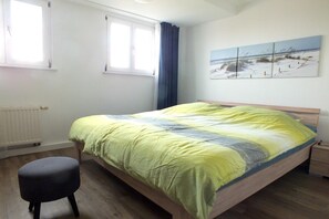 Une chambre à coucher comprenant un lit, une table de chevet, une fenêtre avec des rideaux, un radiateur et une décoration murale sur le thème de la plage.