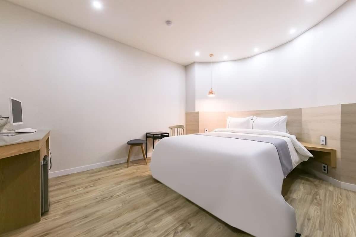 Une chambre d’hôtel moderne avec un lit aux lignes courbes, un bureau avec un ordinateur, un petit tabouret et une table de chevet avec une lampe.