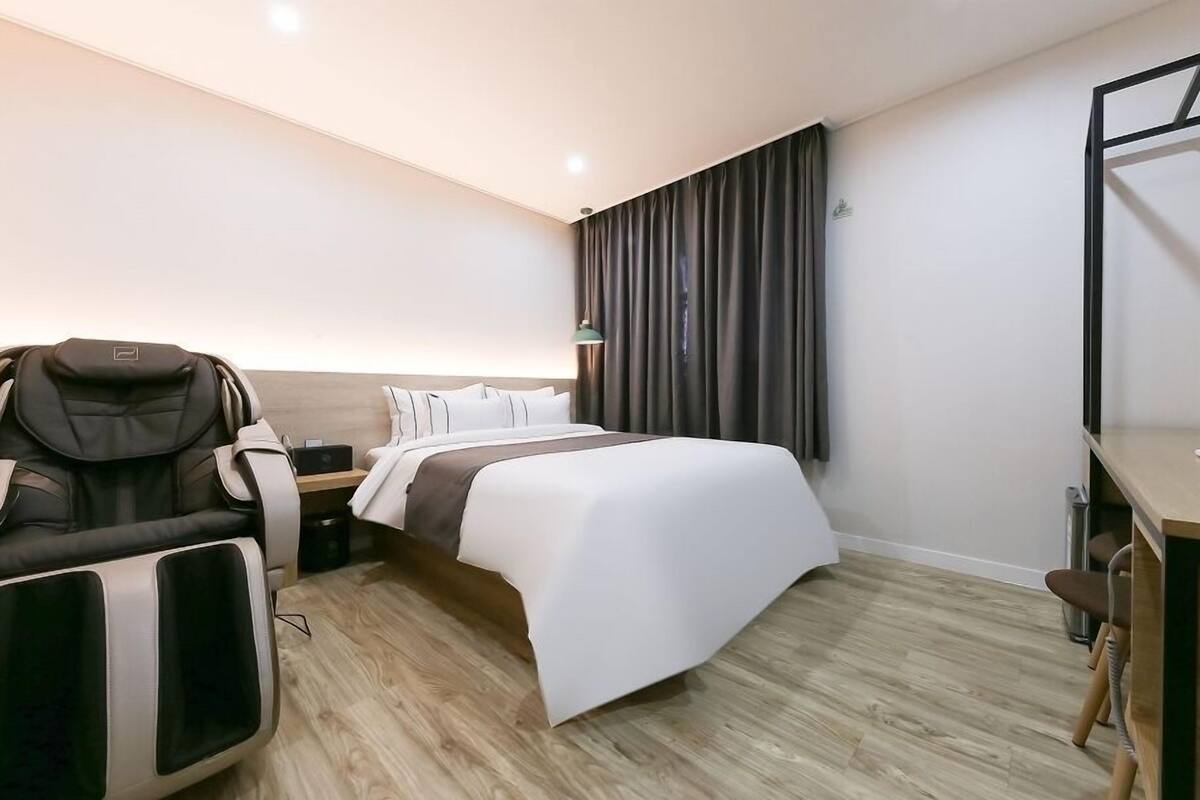 Une chambre d’hôtel équipée d’un lit, d’un fauteuil massant, d’un bureau et d’une grande fenêtre avec des rideaux.