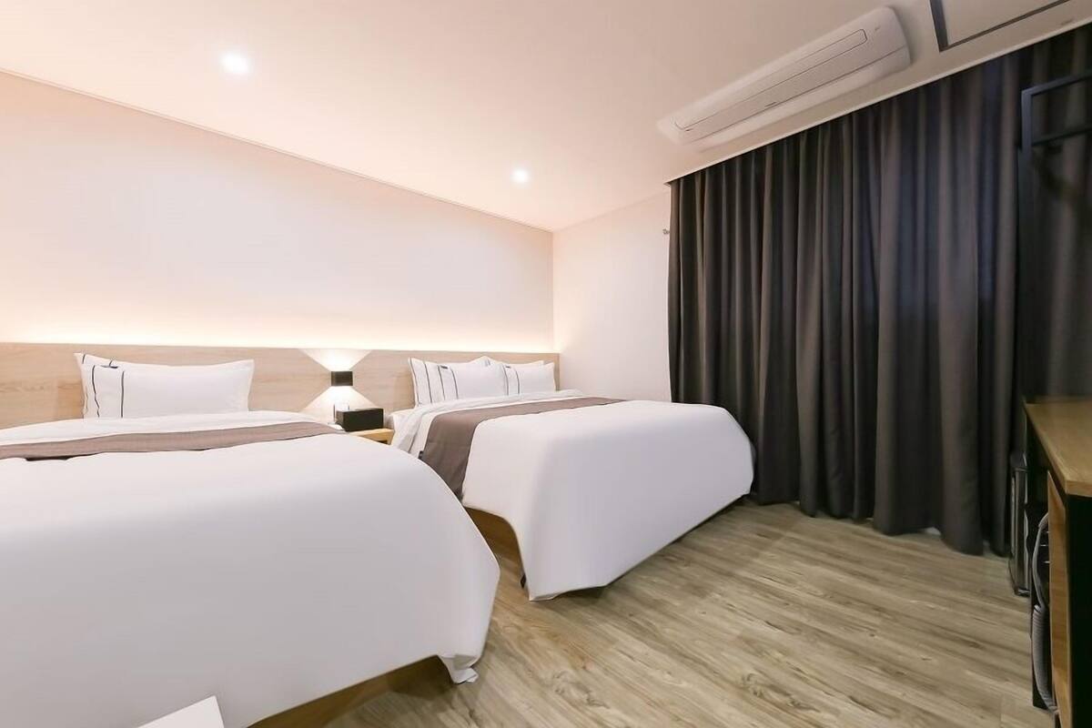 Une chambre d’hôtel avec deux lits, un bureau, une chaise, une lampe et une grande fenêtre avec des rideaux.