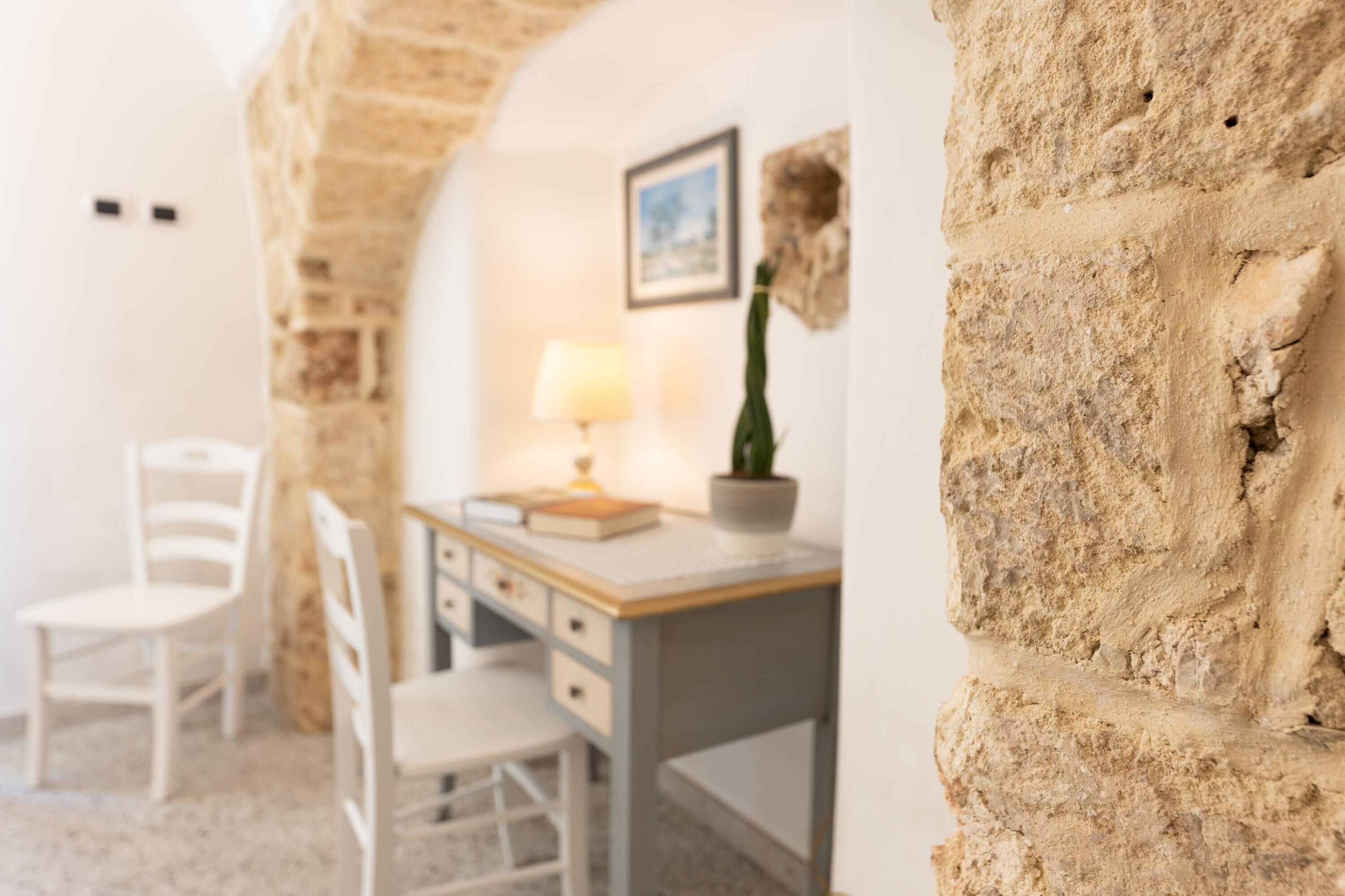 A room with a stone wall, a desk with a lamp, and two chairs.