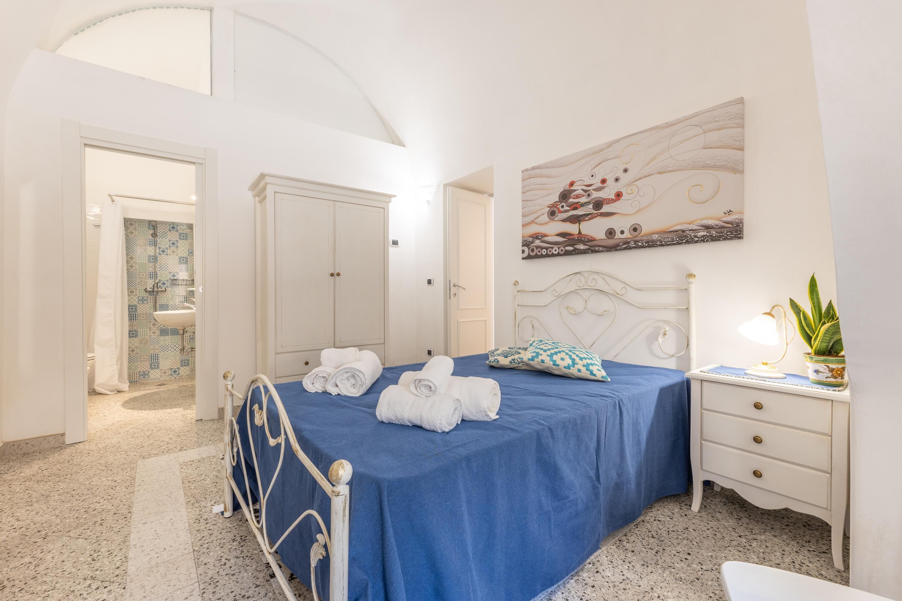 A bedroom with a bed, a nightstand, a desk, and a painting on the wall.