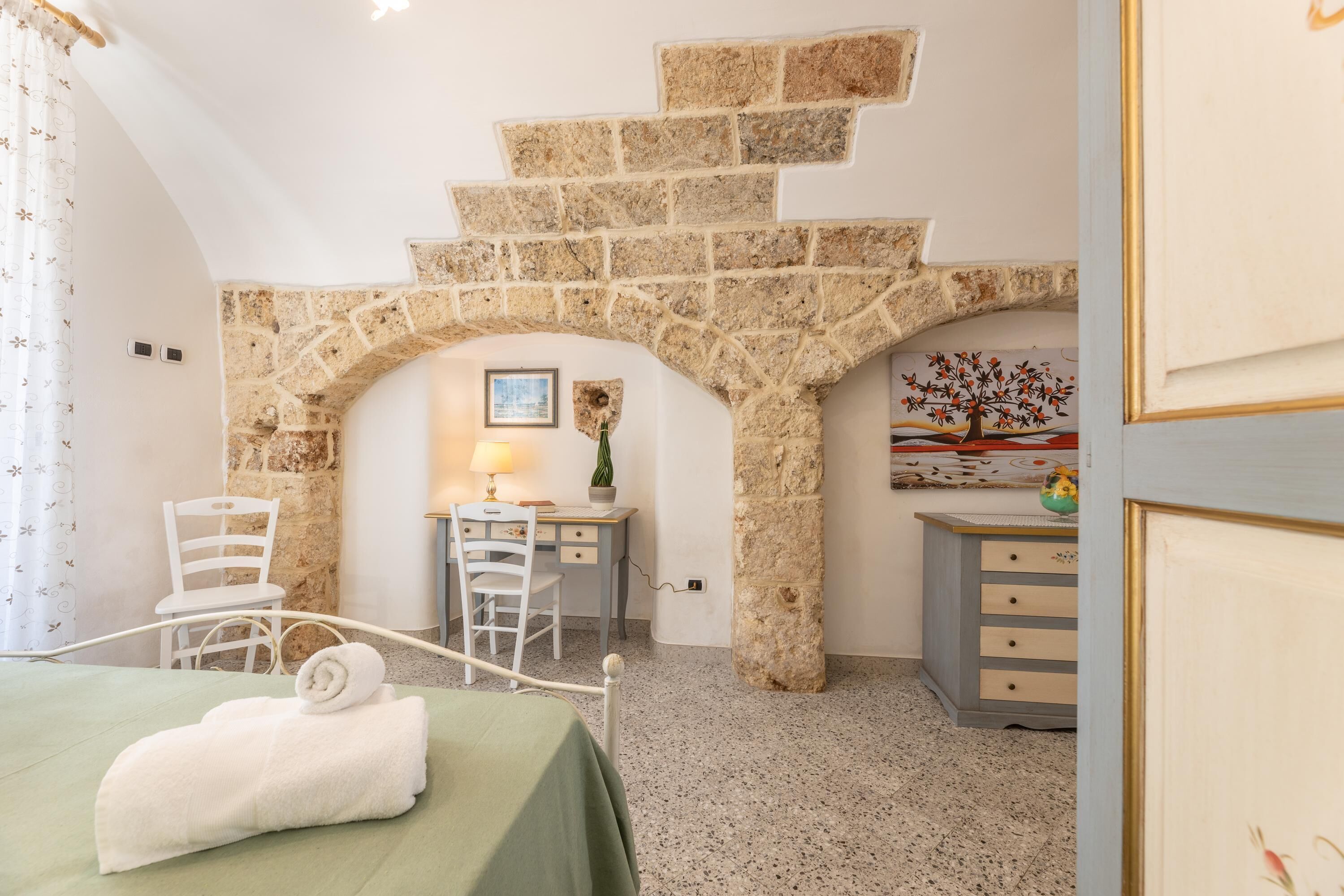 A bedroom with a stone archway, a desk with a lamp, a chair, a bed with a green bedspread, and a dresser.