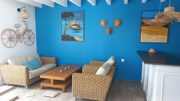 A room with a blue wall, wicker furniture, a wooden table, and a painting of a landscape.