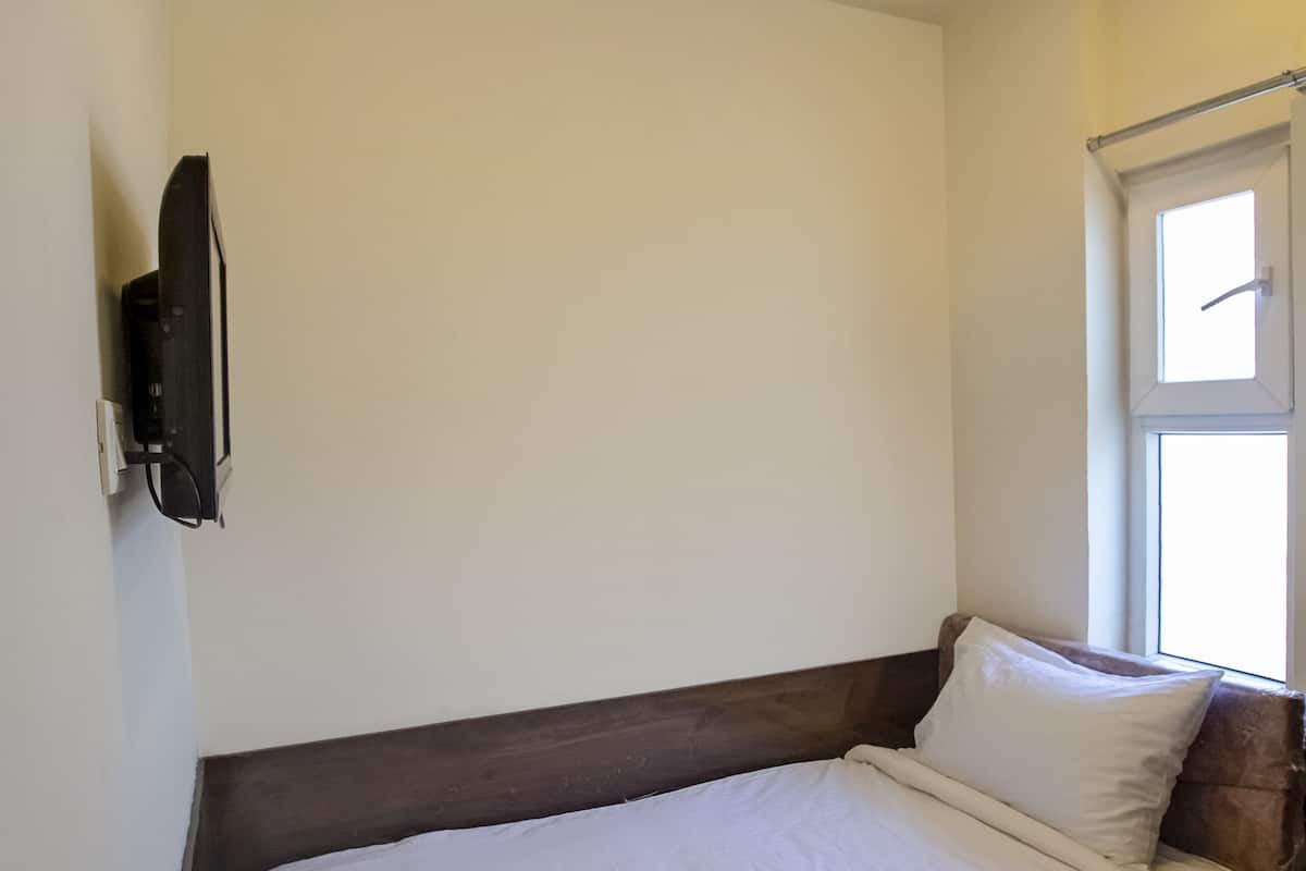 A single bed with a wooden headboard, a ceiling fan, a mounted television, and a window with a view of the outside.