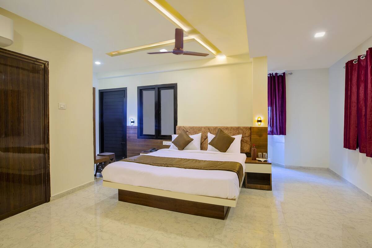A hotel room with a large bed, a ceiling fan, a bedside table with a lamp, and a view of the hallway.