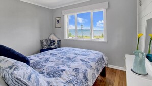 Ein Schlafzimmer mit großem Fenster, einem Bett mit floraler Bettwäsche, einem Nachttisch und einer Vase mit Blumen.
