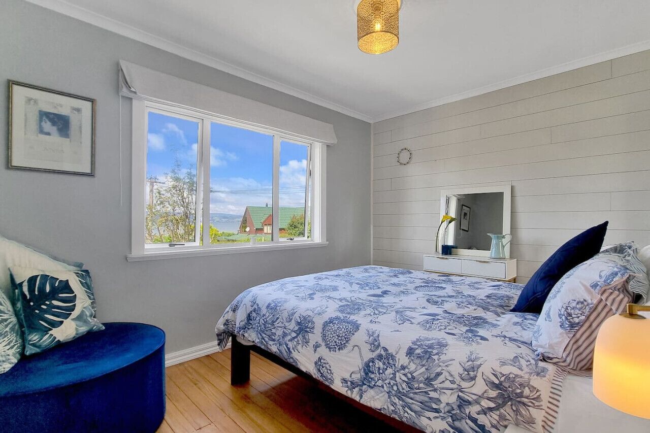 A bedroom with a large window, a bed with floral bedding, a blue chair, and a bedside table with a lamp.