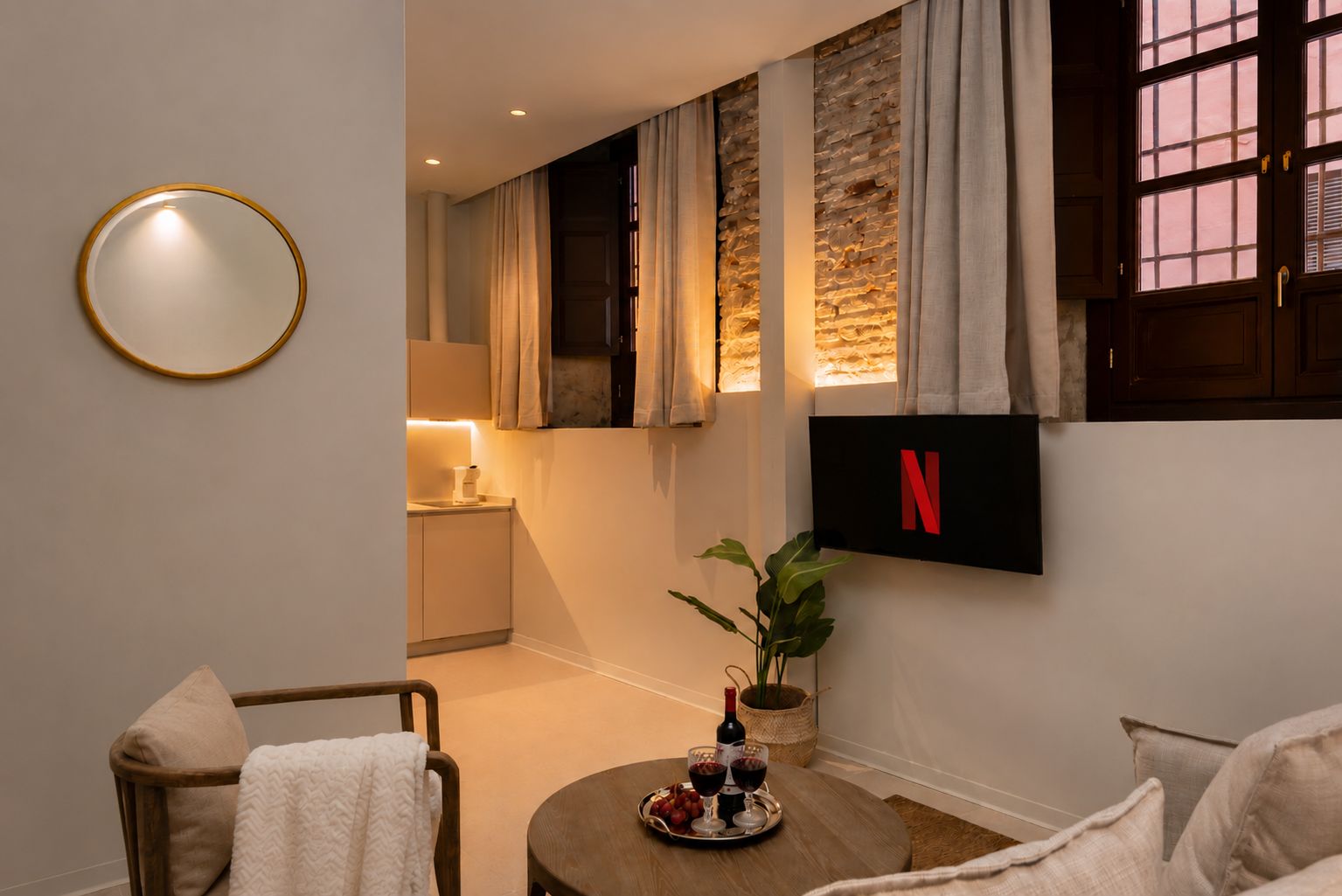 Una sala de estar moderna con un televisor de pantalla plana mostrando el logotipo de Netflix, un espejo redondo, una mesita con una botella de vino y copas, y un rincón de descanso acogedor con un sillón y un sofá.