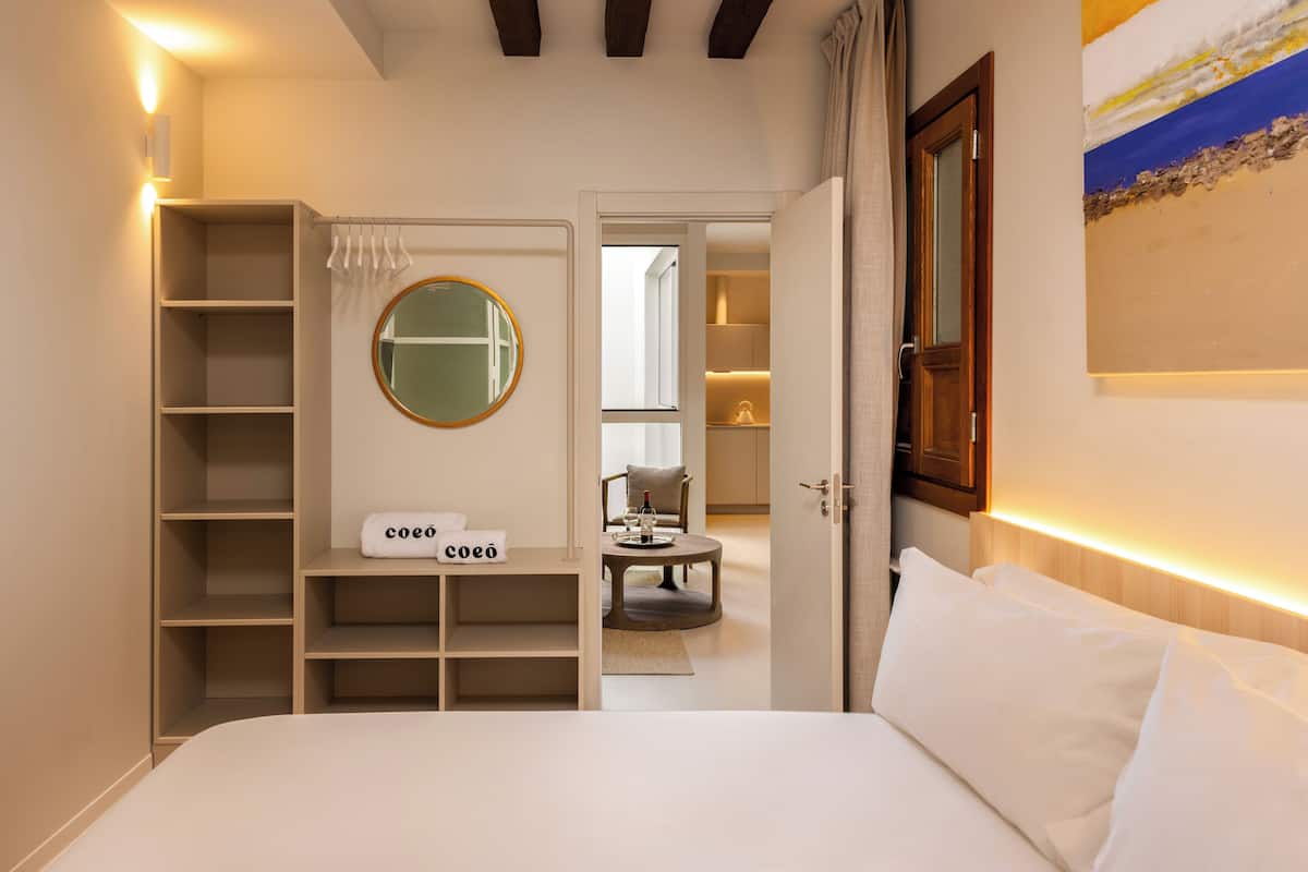 A bedroom with a bed, a bedside table, a mirror, a wardrobe, and a door leading to another room.