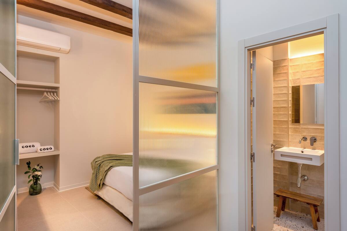 A bedroom with a bed, a bedside table, a small bedside plant, a closet with hanging clothes, and a bathroom with a sink and mirror.