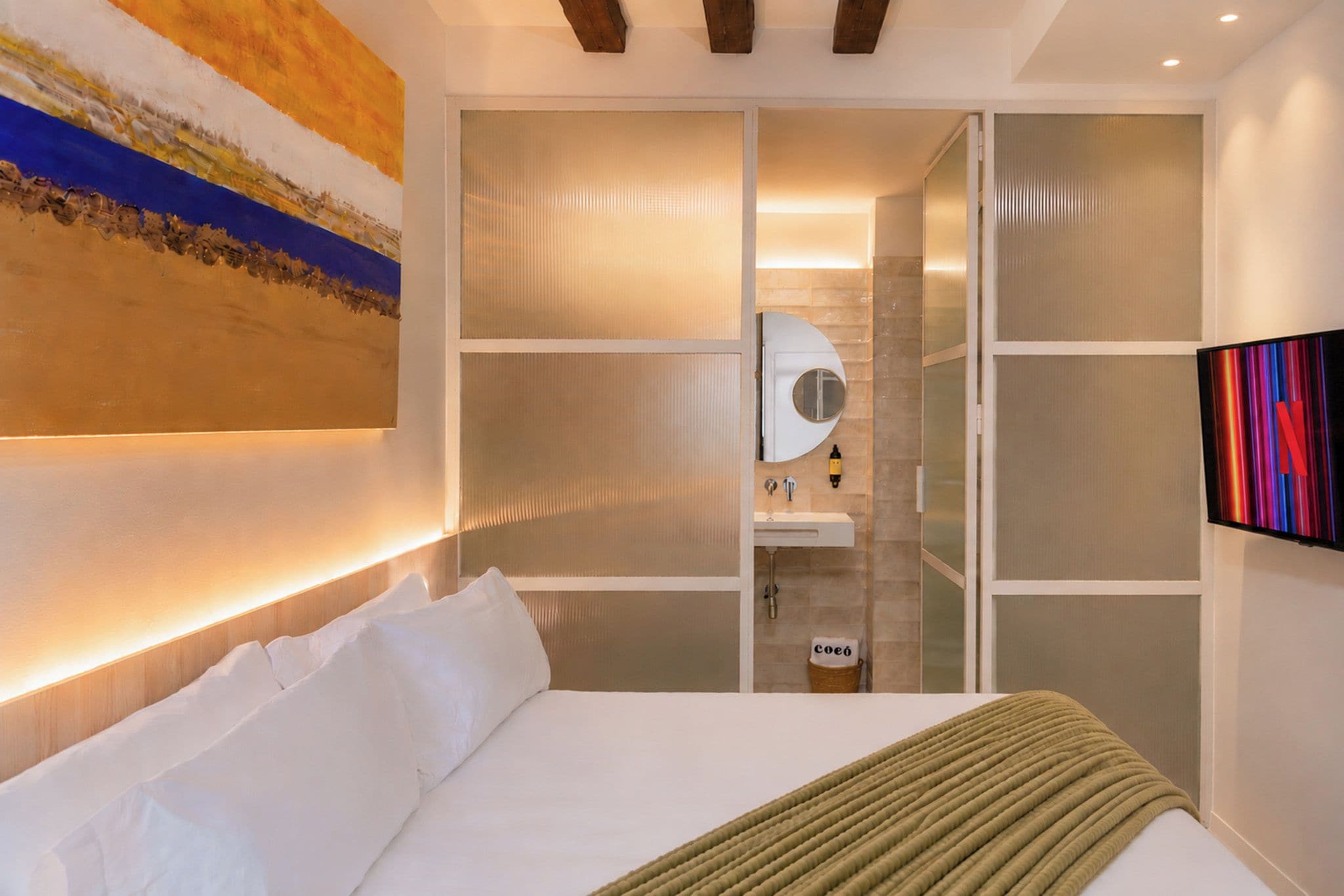 Ein modernes Hotelzimmer mit Bett, einem großen Gemälde, einem Badezimmer mit Spiegel und einem am Wand befestigten Fernseher.