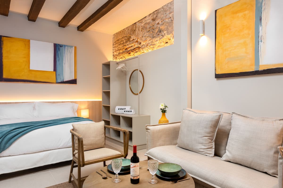 A modern hotel room with a bed, a sofa, a wooden chair, a small table with a bottle and glasses, a mirror, and artwork on the walls.