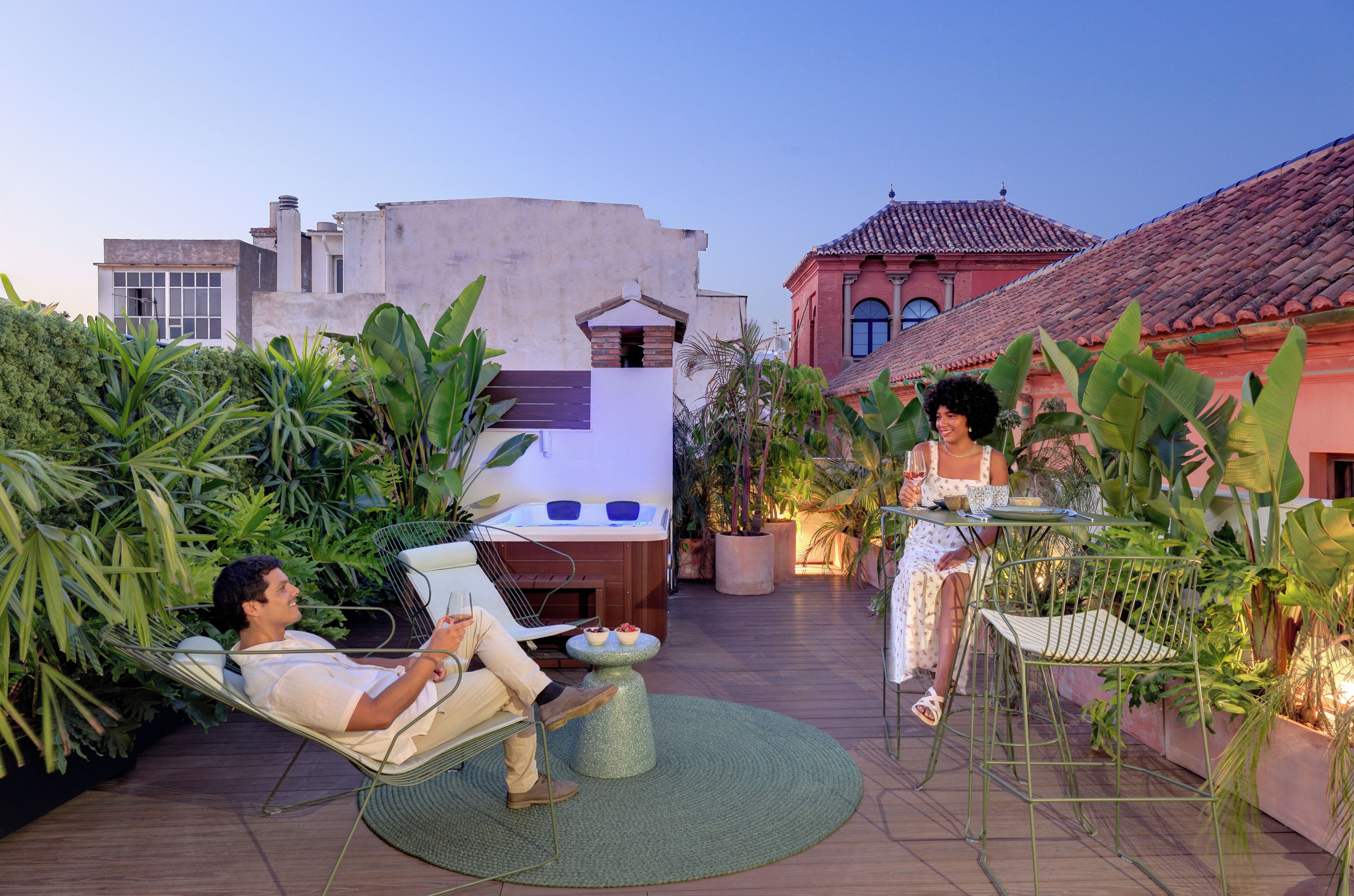 Un homme se détend dans un fauteuil, un ordinateur portable posé sur ses genoux, tandis qu’une femme est assise à une table, un verre à la main, entourée d’une végétation luxuriante et d’un jacuzzi dans une cour.