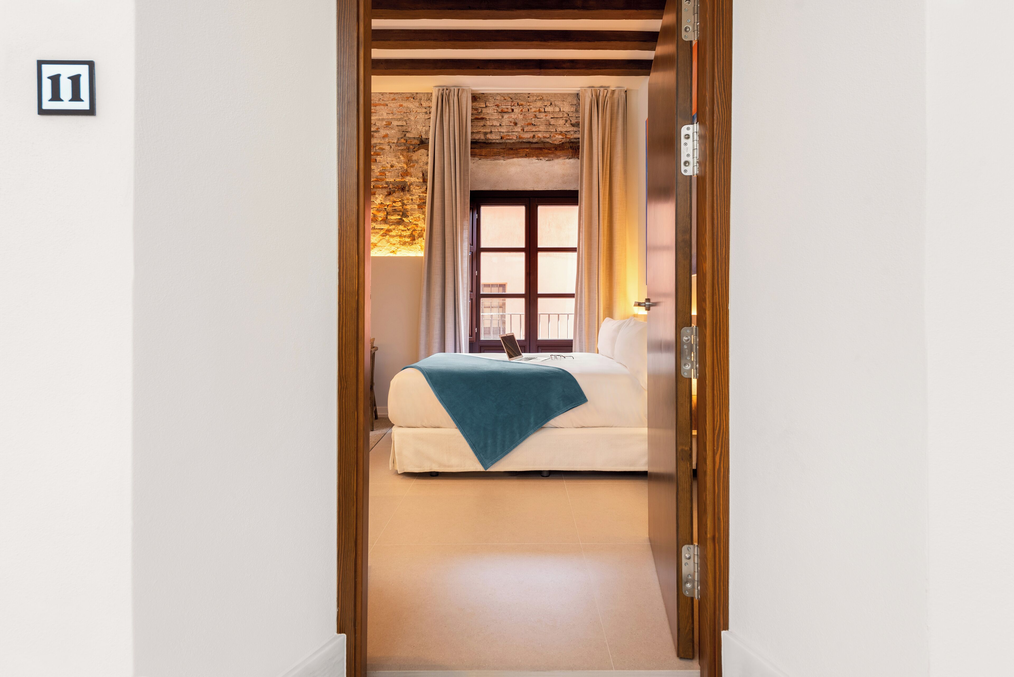 Quarto de hotel com cama, janela com cortinas, teto de madeira e o número 11 na parede.