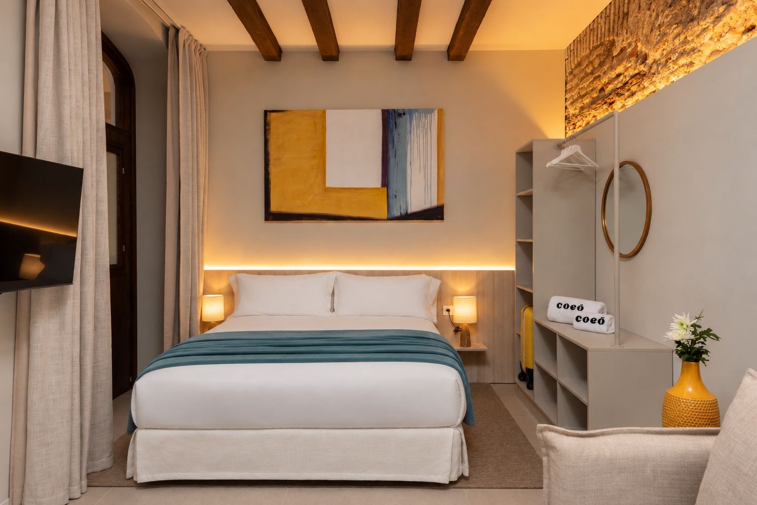 A hotel room with a bed, bedside lamps, a wall-mounted artwork, a shelf with towels, and a chair.