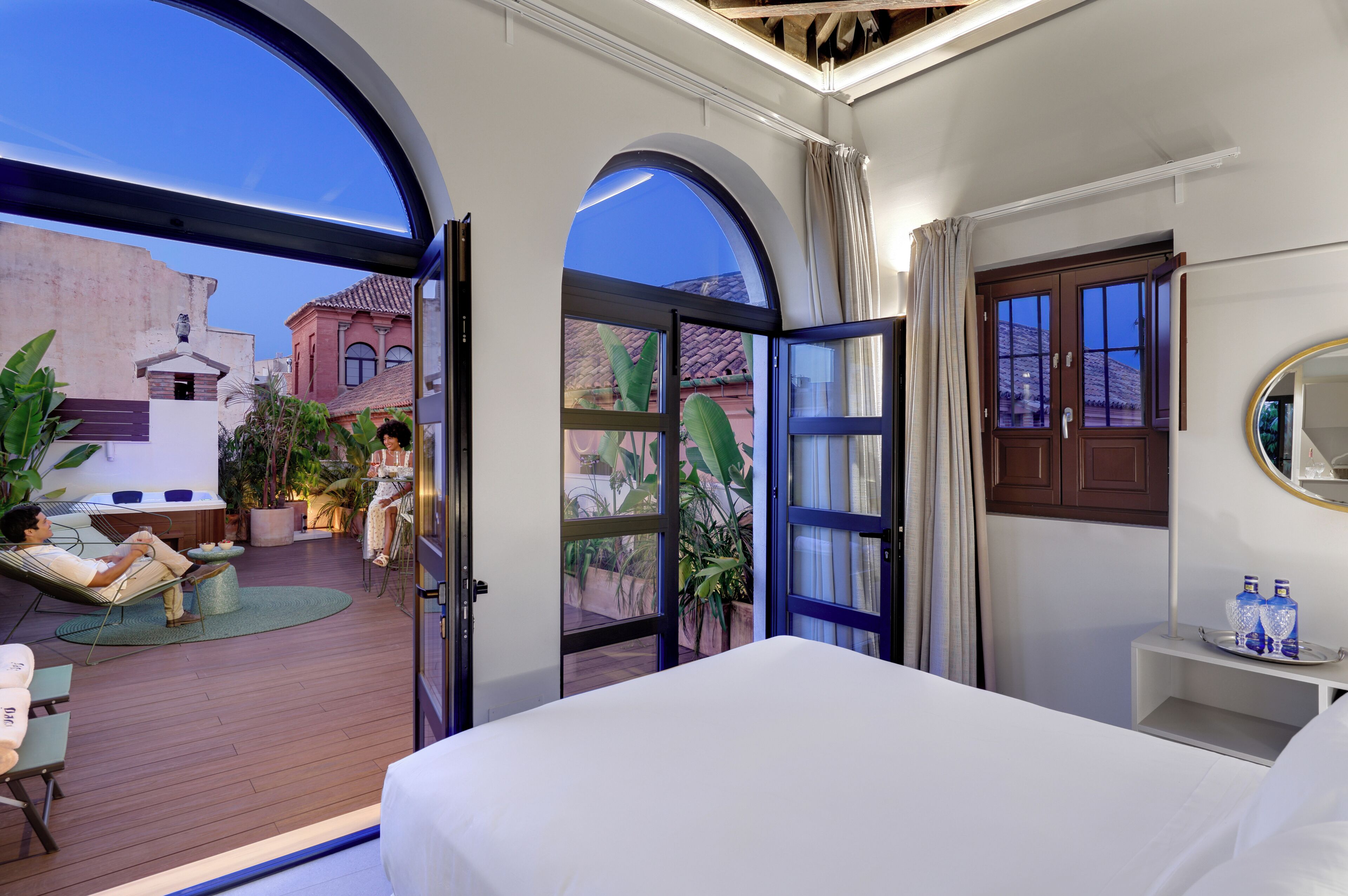 Habitación de hotel con una cama grande, un área de descanso con una silla y una mesa, un balcón pequeño con plantas en macetas y vistas a edificios exteriores.