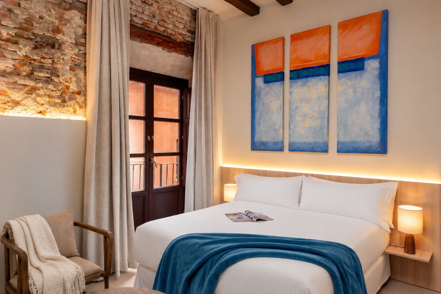 Une chambre d’hôtel avec un mur en briques, un lit avec une literie blanche, un plaid bleu, une chaise en bois et une fenêtre avec des rideaux.