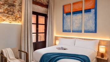 Una habitación de hotel con pared de ladrillo, una cama con ropa blanca, un plaid azul, una silla de madera y una ventana con cortinas.