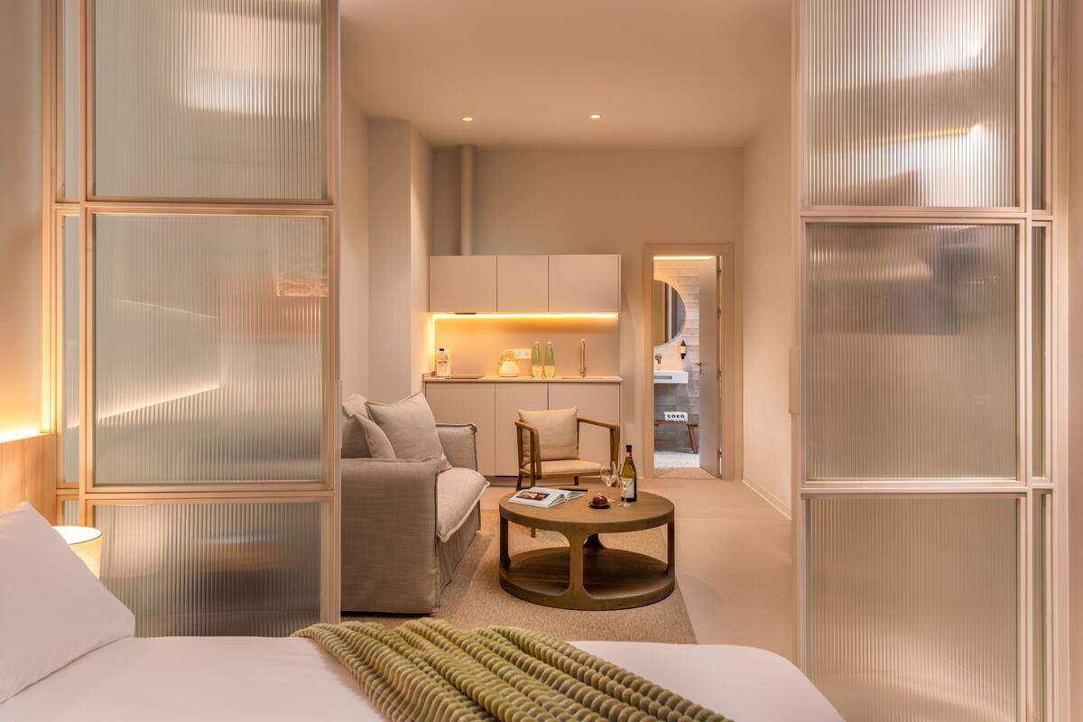 A hotel room with a bed, a round coffee table, a sofa, and a kitchen area in the background.