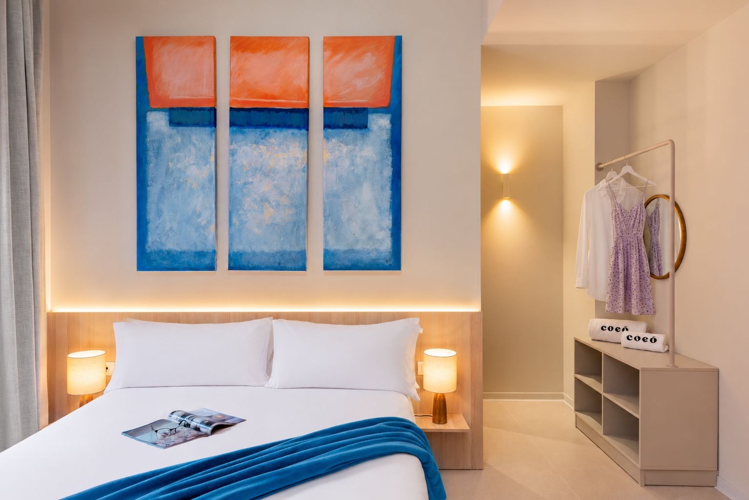 Ein Hotelzimmer mit einem Bett, Nachttischlampen, einer blauen Decke, einer Zeitschrift, einem Gemälde, einem Kleiderschrank und einem Spiegel.