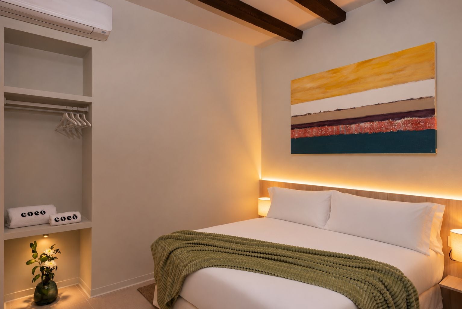 A hotel room with a bed, a bedside lamp, a shelf with towels, and a painting on the wall.