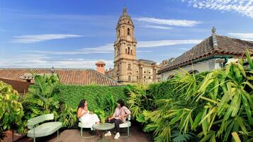 Un jardín en la azotea con dos personas sentadas en una mesa, rodeadas de abundante vegetación y un edificio histórico con una torre de campanas al fondo.