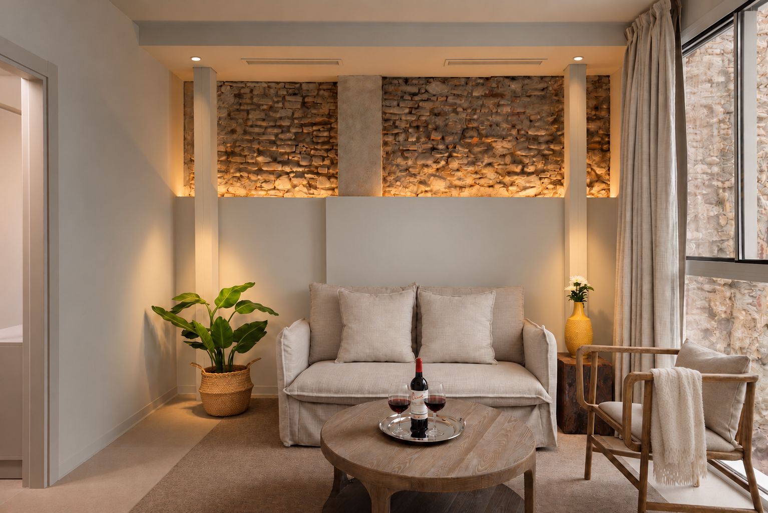 Una sala de estar acogedora con un sofá, una mesita de centro con una botella y vasos, una planta en maceta y una pared de piedra.