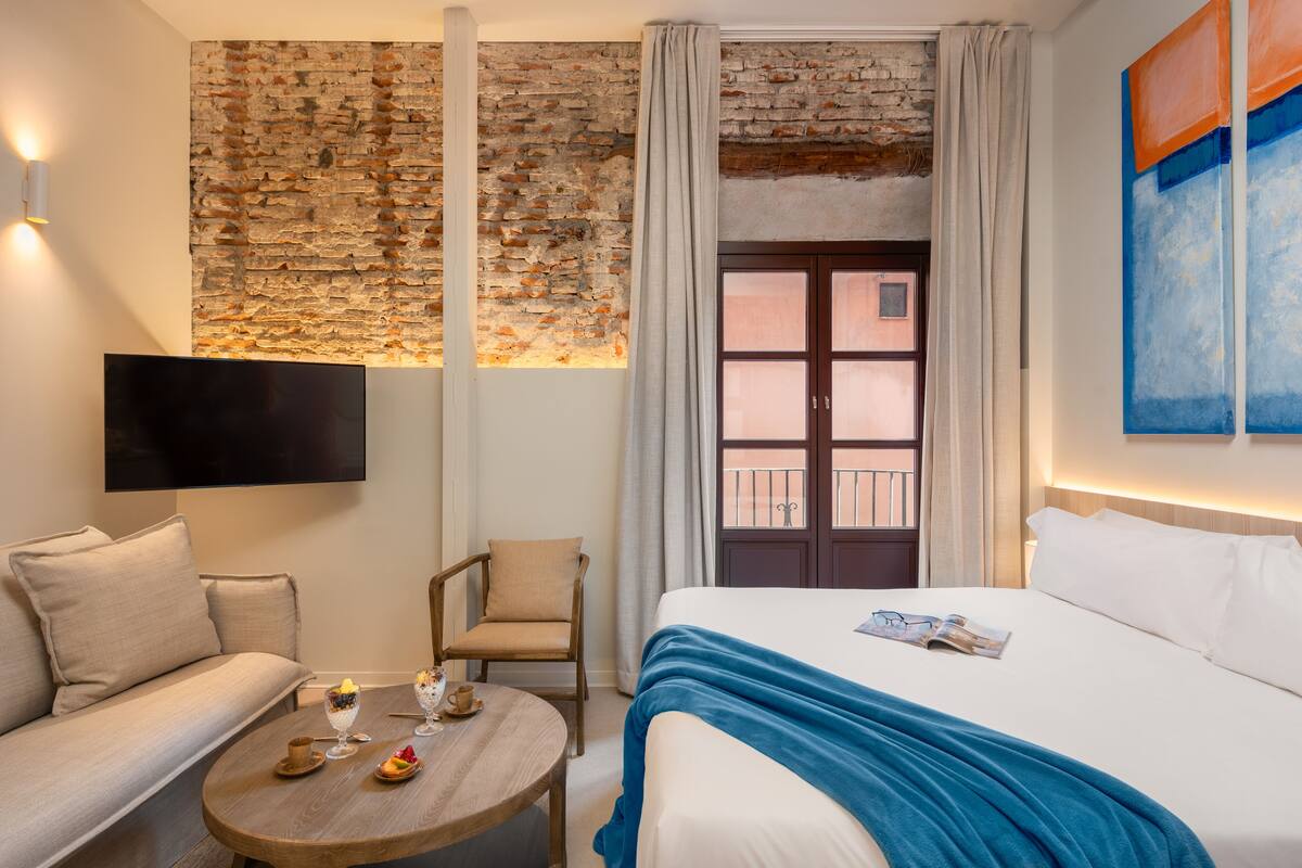 A hotel room with a brick wall, a bed with a blue blanket, a sofa, a chair, a small table with a glass of water and a plate of fruit, and a TV mounted on the wall.