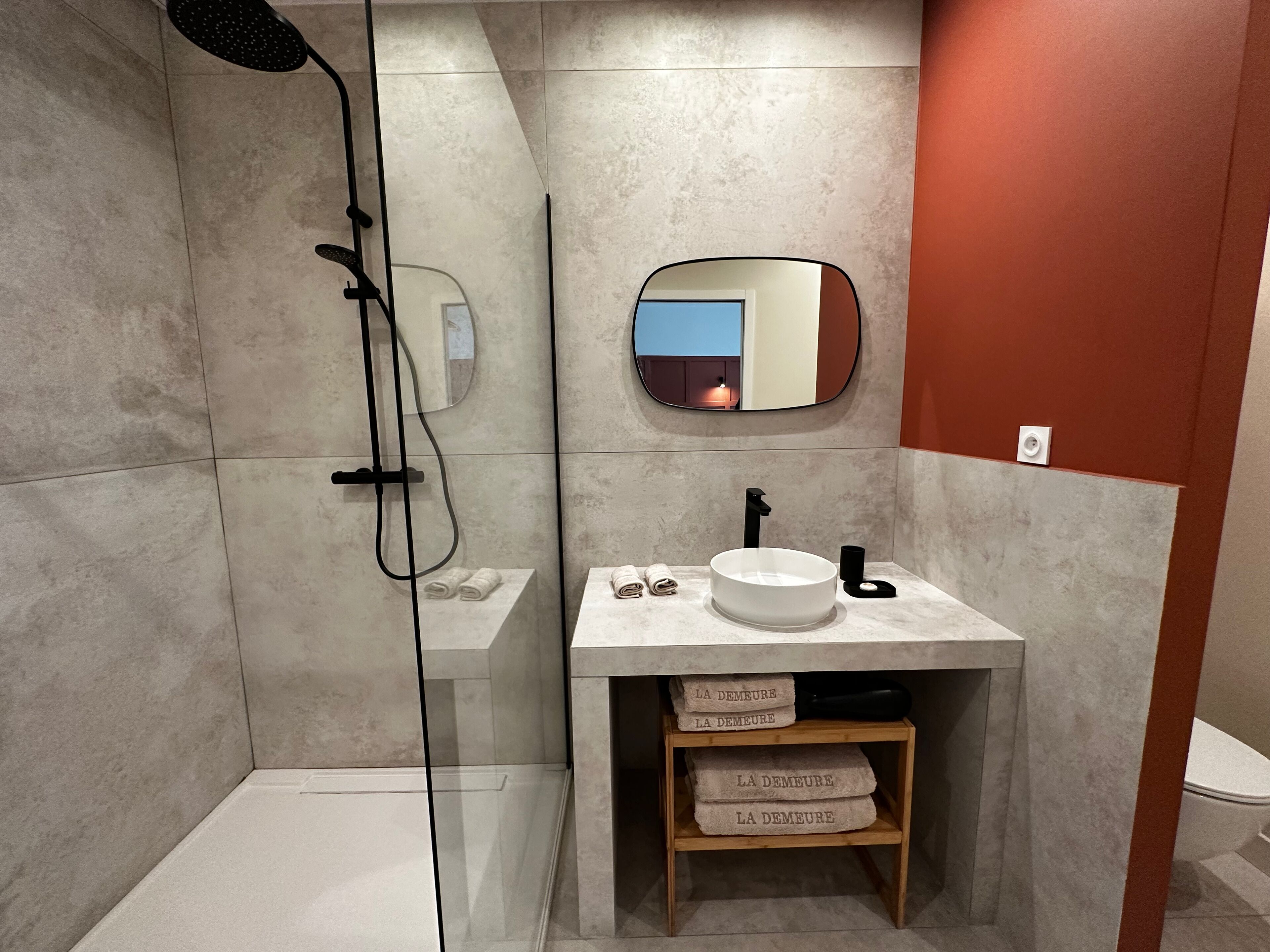 A modern bathroom with a glass-enclosed shower, a white sink with a black faucet, and a mirror above the sink. There are towels stacked on a shelf below the sink.