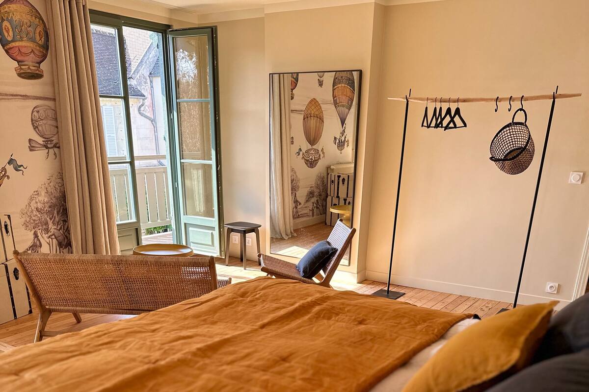 Quarto com uma cama grande, uma cadeira de vime, uma mesa pequena e vista para o exterior através de uma janela.