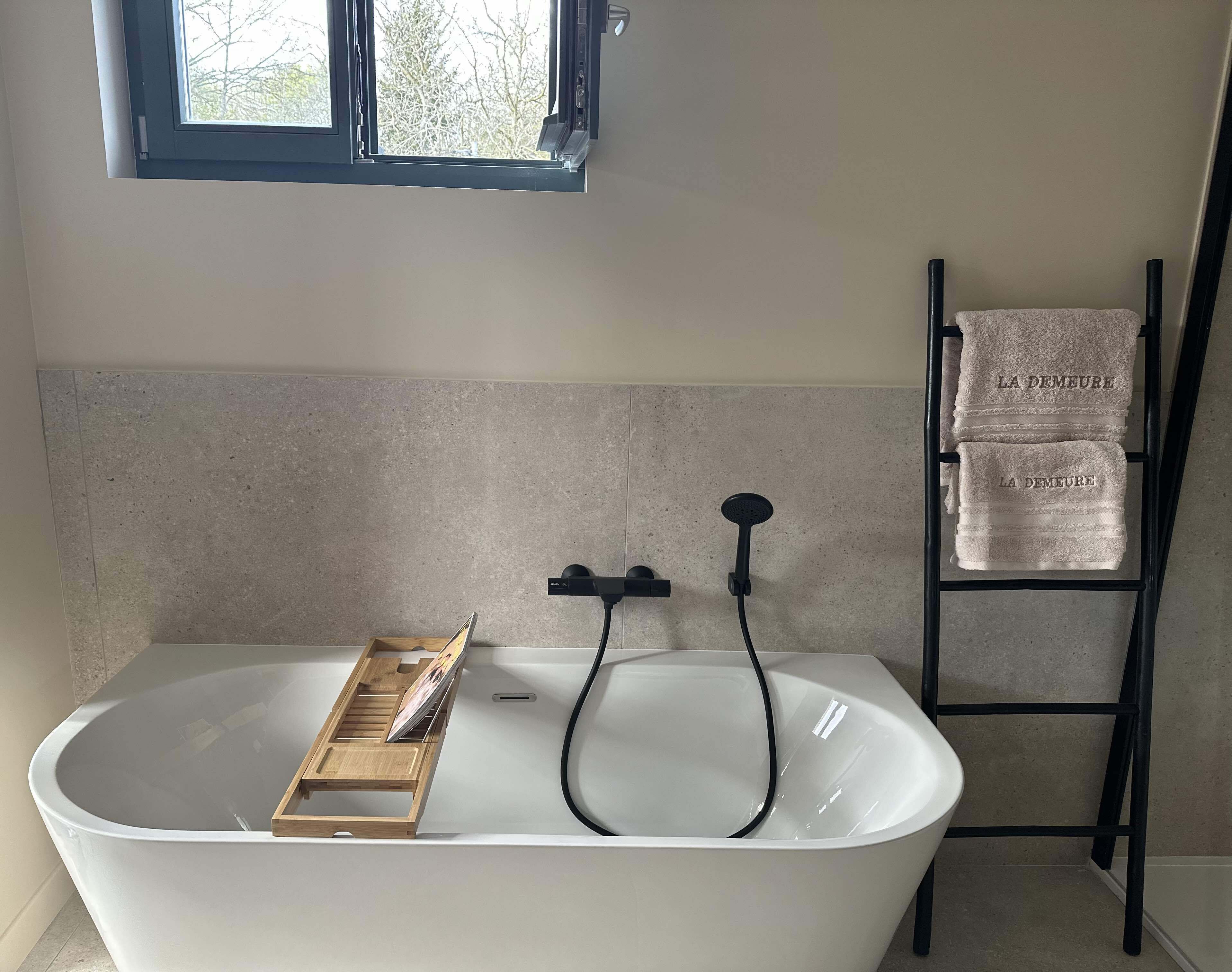 Un baño moderno con una bañera independiente, una bandeja de madera, un cabezal de ducha manual y un perchero con toallas.