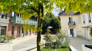 A courtyard with a tree in the foreground, a gravel path, and buildings with balconies and windows. - LA DEMEURE (Précy-sur-Oise)