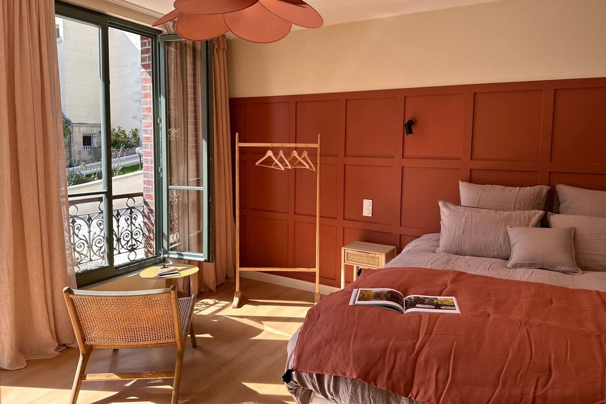 Quarto de hotel com uma cama grande, uma mesinha, uma cadeira, uma janela com cortinas e um lustre.