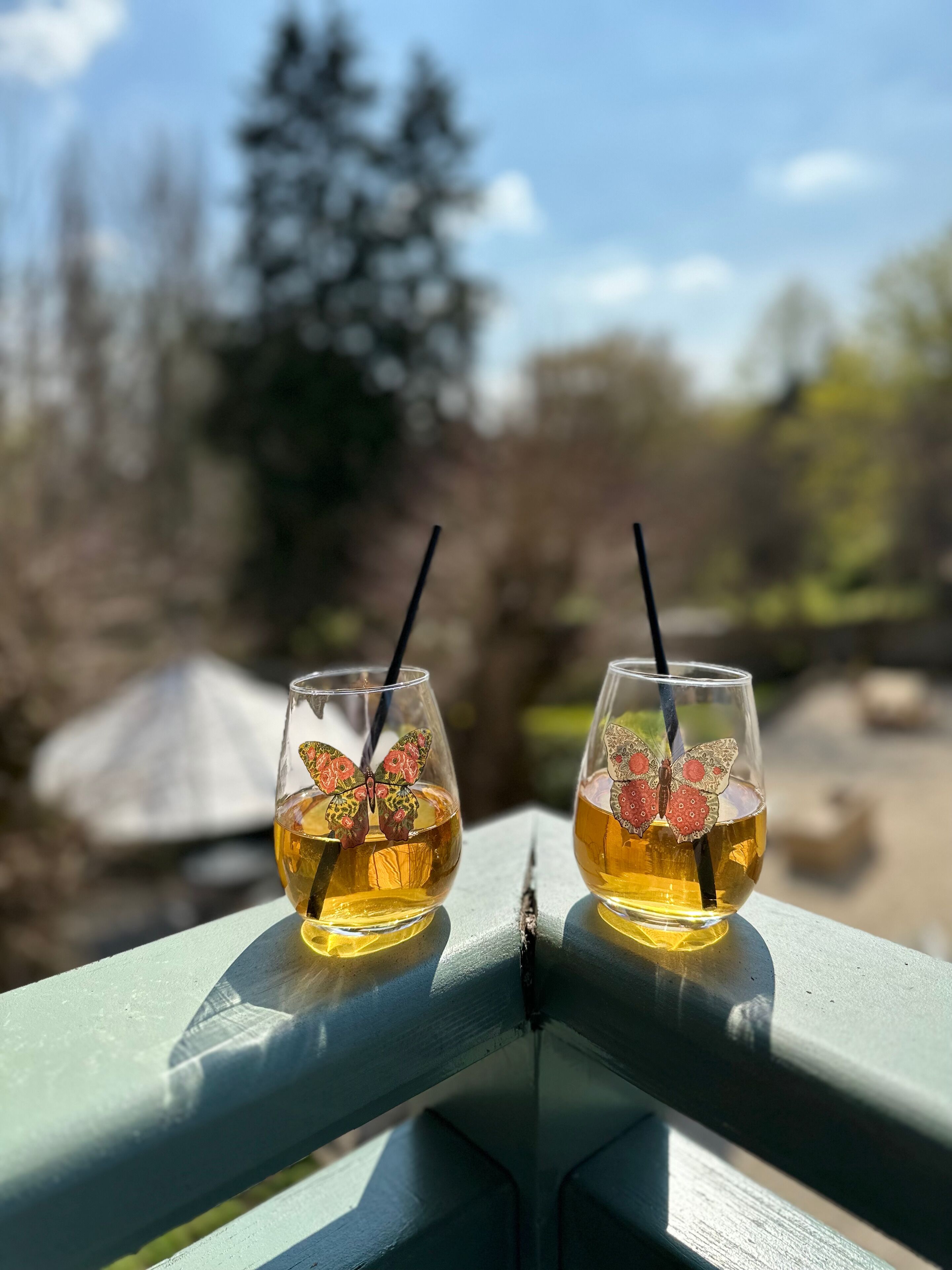 Dos vasos de líquido ámbar con decoraciones de mariposa sobre una barandilla, con un fondo natural desenfocado.