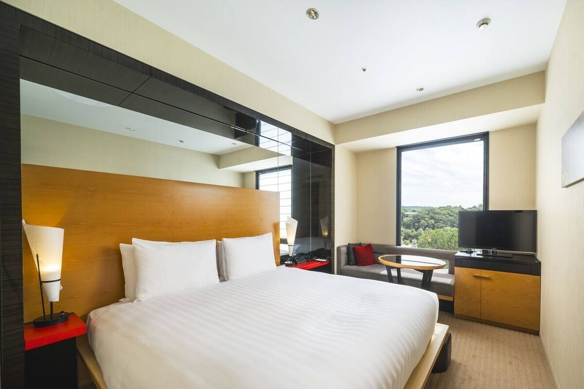 Ein Hotelzimmer mit einem großen Bett, einem Fernseher, einem Schreibtisch und einem Fenster mit Blick.