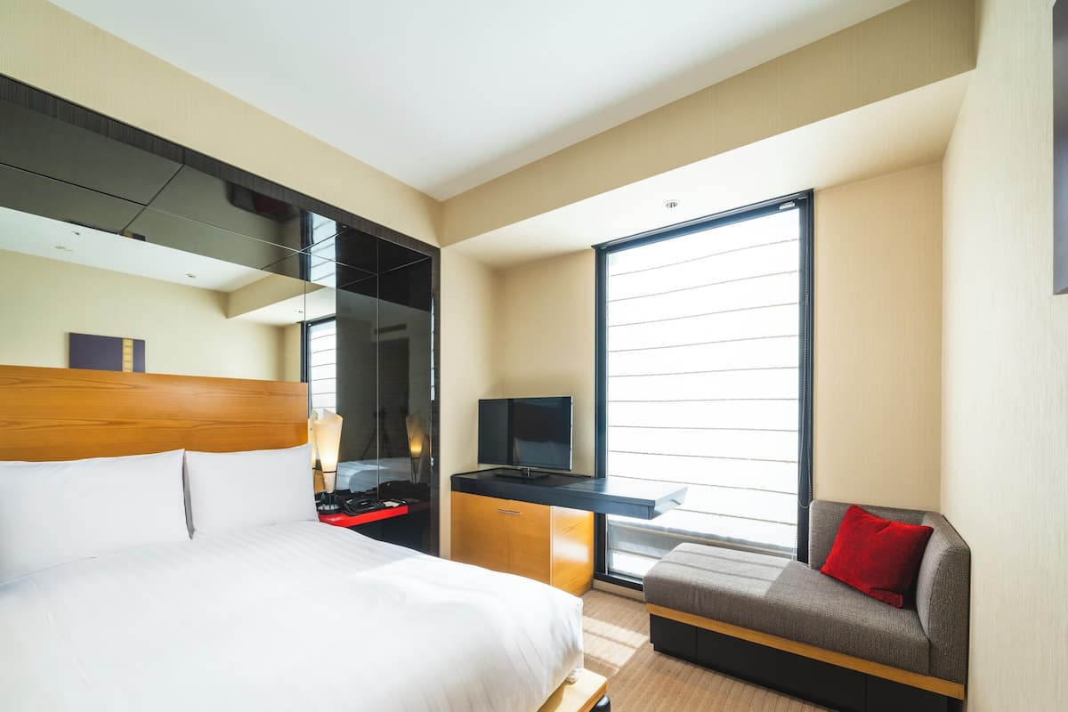Ein Hotelzimmer mit einem Bett, einem Schreibtisch mit Fernseher, einer Couch mit rotem Kissen und einem großen Fenster.