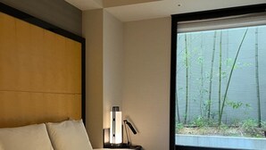 A hotel room with a large bed, a nightstand, a lamp, and a window with a view of bamboo trees. - Koko Hotel Premier Kanazawa Korinbo (Kanazawa)