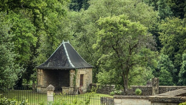 Edifício de pedra com telhado pontiagudo, cercado por vegetação exuberante e uma cerca de metal.