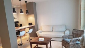 Uma sala de estar moderna com um sofá, uma poltrona de vime, uma mesa de centro e uma área de cozinha com banquetas.